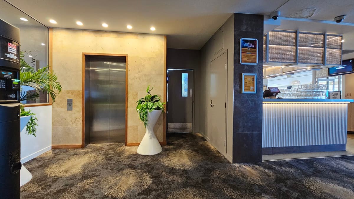 On the left, there is a black vending machine and a potted plant beside a wall. In the middle, there is a stainless steel lift with a beige wall and recessed lighting above it. In the background, there is a small hallway leading to a door. On the right, there is a reception counter with a ribbed white front and illuminated base, along with framed certificates on the wall. The floor is covered in dark, patterned carpet, and the ceiling has recessed lighting.