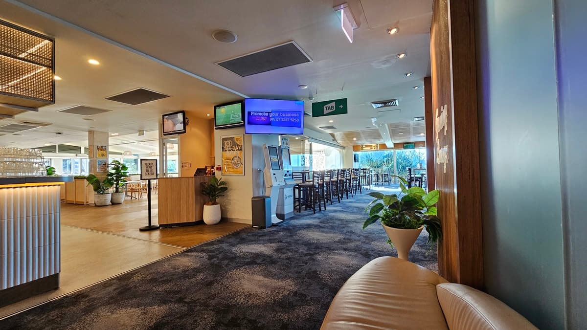 On the left, there is a reception counter with a ribbed white front and a potted plant beside it. In the middle, there is a digital kiosk and a TV screen mounted above it, with a hallway leading to a dining area in the background, featuring tables and chairs. On the right, there is a beige curved sofa and a large potted plant. The floor is covered in dark, patterned carpet, and the ceiling has recessed lighting and air conditioning vents.