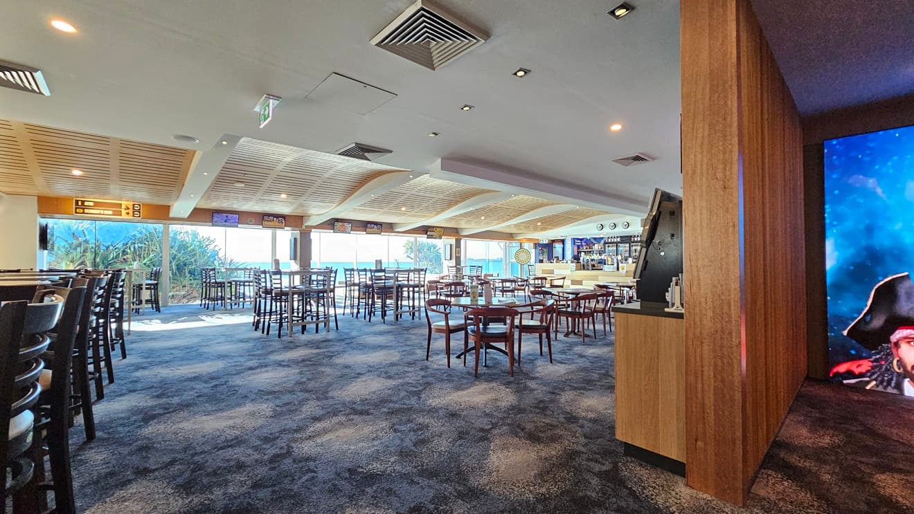 On the left, there are tall tables with high chairs and large windows offering a view of greenery. In the middle, there are multiple round tables with wooden chairs spread across the room, and a wooden ceiling with recessed lighting. In the background, there is a bar area with stools and a wall-mounted TV. On the right, there is a wooden partition wall with a digital display showing a pirate image. The floor is covered in dark, patterned carpet, and the ceiling has recessed lighting .