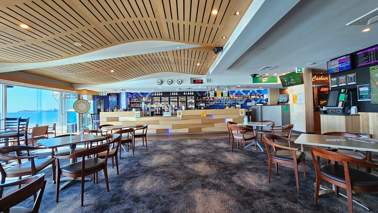 On the left, there are large windows with a view of the sea and a dartboard mounted on the wall. In the middle, there are several wooden tables and chairs on a dark, patterned carpet. In the background, there is a long bar counter with stools, various bottles, and a digital clock above it. On the right, there are more tables and chairs, a wall-mounted TV, and a gaming machine. The ceiling is wooden with a wave-like design and recessed lighting.