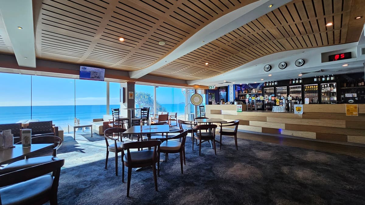 On the left, there are large windows with a view of the sea, a dartboard on the wall, and a few tables with chairs. In the middle, there are several round wooden tables with chairs on a dark, patterned carpet. In the background, there is a long bar counter with stools, various bottles, and a digital clock above it. On the right, there are more tables and chairs, a wall-mounted TV, and a gaming machine. The ceiling is wooden with a wave-like design and recessed lighting.