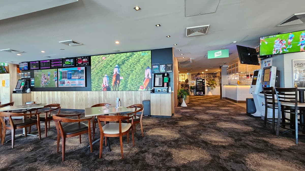 On the left, there are several wooden tables with chairs and large wall-mounted screens displaying various images and information. In the middle, there is a hallway with a dark, patterned carpet leading to a bar area in the background, featuring stools and illuminated shelves with bottles. On the right, there is a gaming machine, a TV screen mounted above it, and a high table with stools. The ceiling has recessed lighting and air conditioning vents.