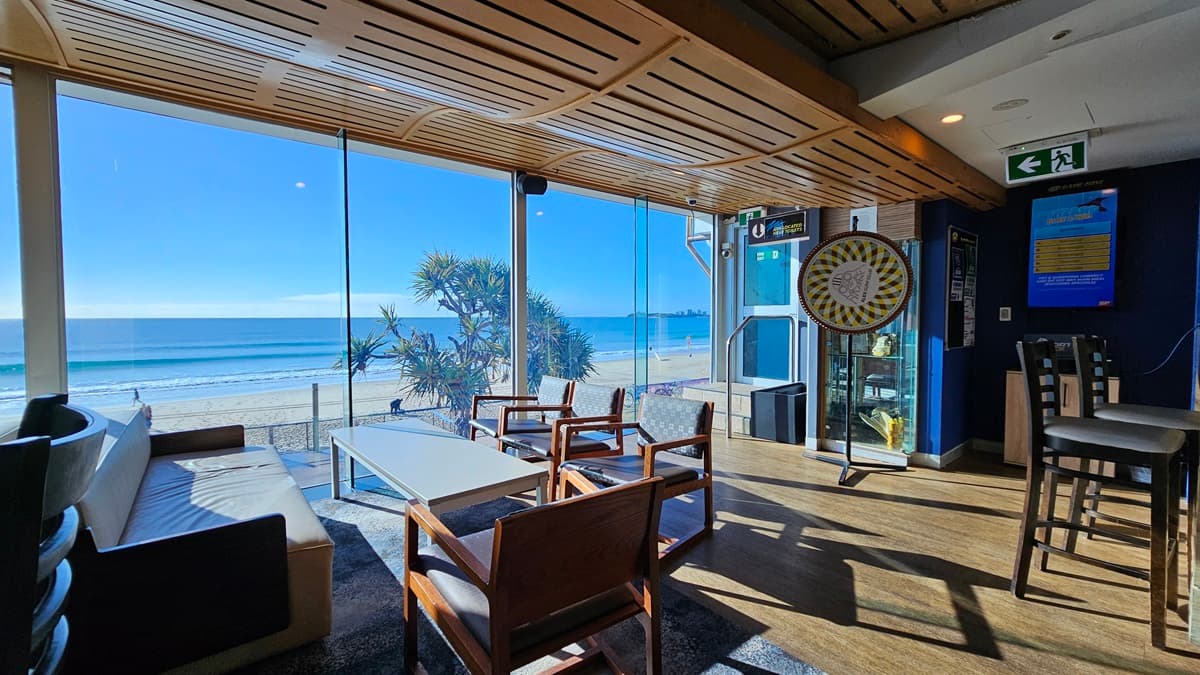On the left, there is a black sofa with a white cushion. In the middle, there are wooden chairs and a white coffee table on a dark rug, with large windows offering a view of the beach and sea. In the background, there is a dartboard mounted on a blue wall and a vending machine. On the right, there are high tables with stools and an emergency exit sign above. The floor is wooden, and the ceiling has a wooden slat design with recessed lighting.