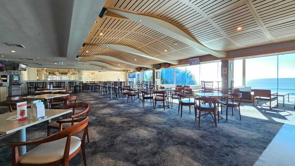On the left, there are several wooden tables with chairs and a few wall-mounted screens. In the middle, there are more wooden tables and chairs on a dark, patterned carpet, with large windows offering a view of the sea. In the background, there is a long bar counter with stools and illuminated shelves. On the right, there are additional tables and chairs, and a lounge area with sofas. The ceiling has a wave-like wooden slat design with recessed lighting.