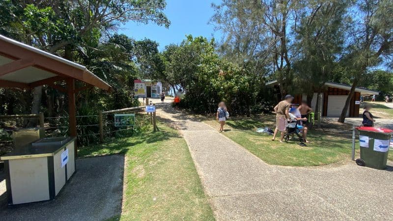 On the left, a covered barbecue area with a stone bench and a nearby fence sits next to grassy ground. On the right, a group of people stand near a small wooden building with bins on the path's edge. In the middle, a paved path leads towards a gap in the trees, revealing beach access signs and a glimpse of sandy ground in the background. The floor consists of a mix of concrete paving, grass, and patches of sand. There is no visible ceiling.