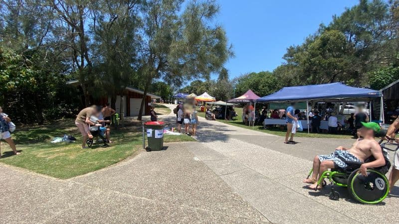 On the left, a covered barbecue area with a metal bench sits beside a railing and green grass. On the right, a group of people stand near a bin, a stroller, and a small wooden building partially visible behind trees. In the middle, a paved path leads upwards through dense greenery towards beach access signs and a glimpse of sandy ground at the path's end. The floor consists of concrete, grass, and sand.