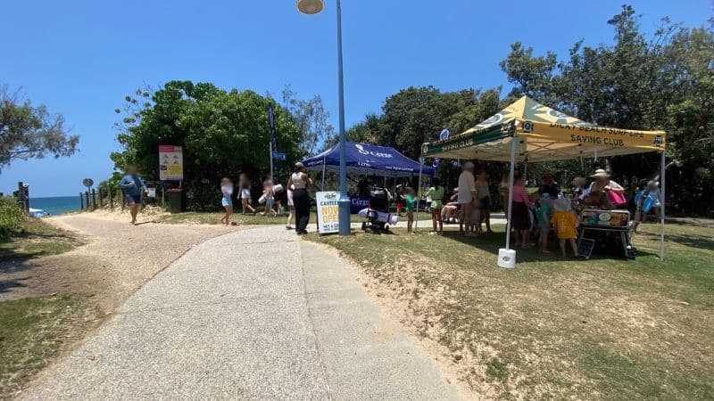 On the left, a grassy area bordered by dense trees contains a yellow warning sign and a bin near a blue "Toilets" signpost. On the right, a paved pathway curves through grass and trees, with several people walking in groups. In the middle, a dark blue gazebo branded with white text is set up on the grass, with promotional flags on either side. The foreground includes a mix of gravel and grass, with a partly visible black railing.