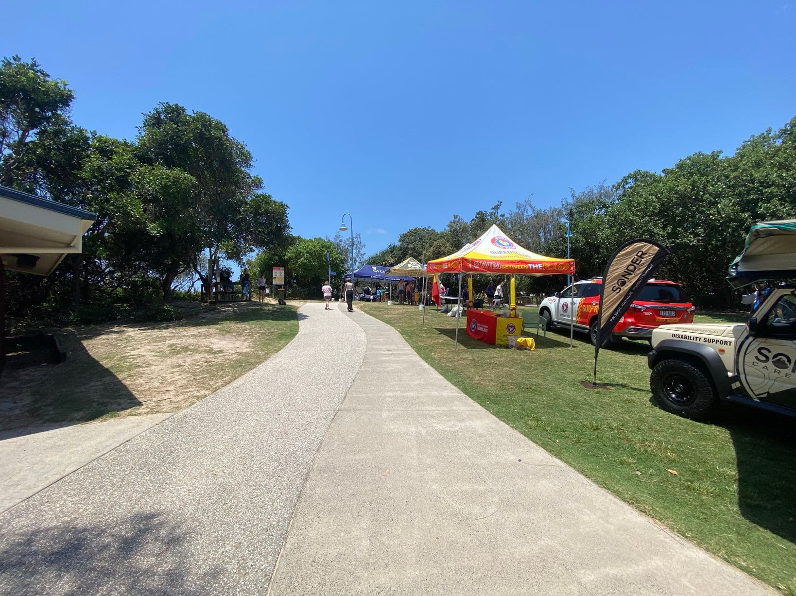 On the left, a sandy path bordered by grass and a wooden fence leads towards the beach, with a signboard and people walking nearby. On the right, two gazebos, one blue and one yellow, are set up on grass with groups of people gathered underneath. In the middle, a wide paved path runs toward the background, flanked by grass and small patches of sand. The foreground features the textured paved path and some scattered patches of dirt. A tall lamppost stands centrally.