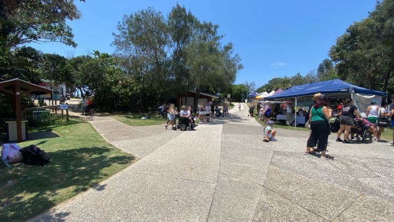 On the left, a grassy area with a small wooden shelter and a bin is surrounded by shrubs and trees. On the right, blue and white gazebos are set up on grass, with people gathered underneath and a pram nearby. In the middle, a wide paved path extends towards the background, leading to a small building and further greenery. The foreground consists of textured concrete paving, grass, and scattered shadows.