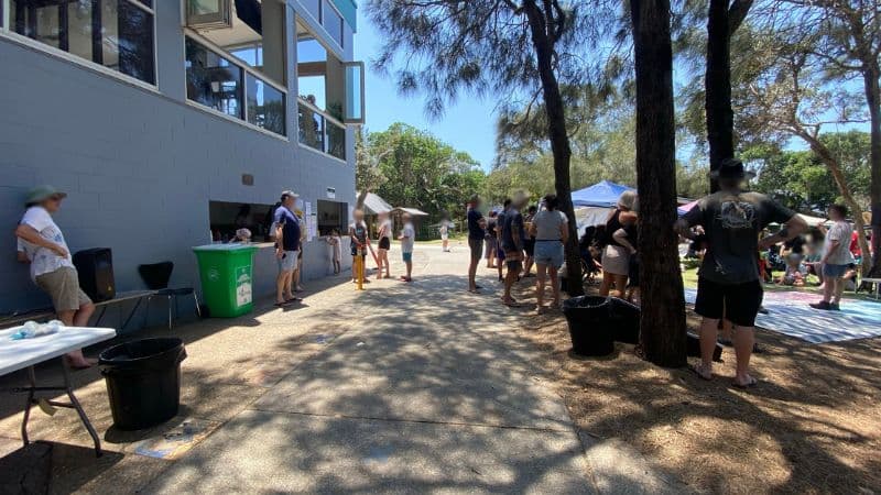 On the left, a light grey building with open service windows has people standing nearby, next to green and black bins and a white folding table. On the right, a group of people are gathered around gazebos and seated on mats under tall trees. In the middle, a paved pathway stretches forward, with scattered shadows and tree trunks along its sides. The floor consists of textured concrete, dirt, and pine straw.