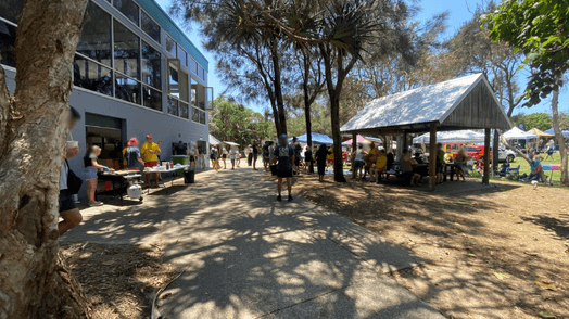 On the left, a grey building with large glass panels has people gathered beside tables holding items, with bins and vegetation nearby. On the right, a wooden pavilion with a metal roof shelters seated people, while open grass and market-style gazebos with more people are visible further back. In the middle, a paved path curves through the scene, bordered by trees casting dappled shadows. The floor includes concrete, dirt, and patches of grass.