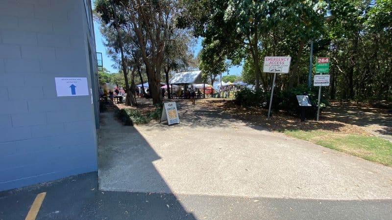 On the left, a blue-grey brick building has a white sign with a blue arrow attached to its wall. On the right, signs reading “Emergency Access” and “264” are mounted on poles near bushes, with a black information board positioned below. In the middle, a wide concrete path leads to a shaded area under trees featuring gazebos and people gathered. The foreground consists of concrete and asphalt, with greenery visible along the edges.