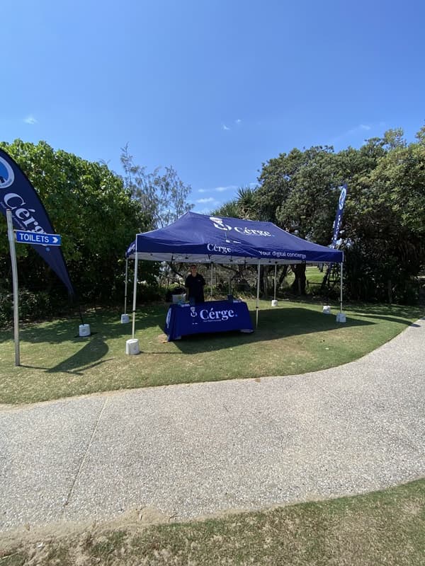On the left, a vertical blue flag reading "Toilets" stands on grass next to bushes and trees. On the right, a cluster of dense greenery surrounds another blue flag. In the middle, a blue gazebo with "Cérge" branding on the roof and tablecloth sits on a lawn, partially shaded by trees. A person stands under the gazebo behind a table. The foreground features a textured, light-coloured curved concrete path bordered by grass.