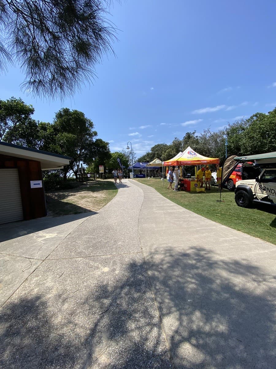 On the left, a small brown building with a closed roller door is partially in shade, next to sandy ground and grass bordered by trees. On the right, a bright yellow and orange gazebo with tables and items for display is set up on grass, alongside a parked vehicle with a partially extended awning. In the middle, a curved, textured concrete path extends into the distance, surrounded by grass and trees. The foreground includes light and shadow patterns on the concrete.