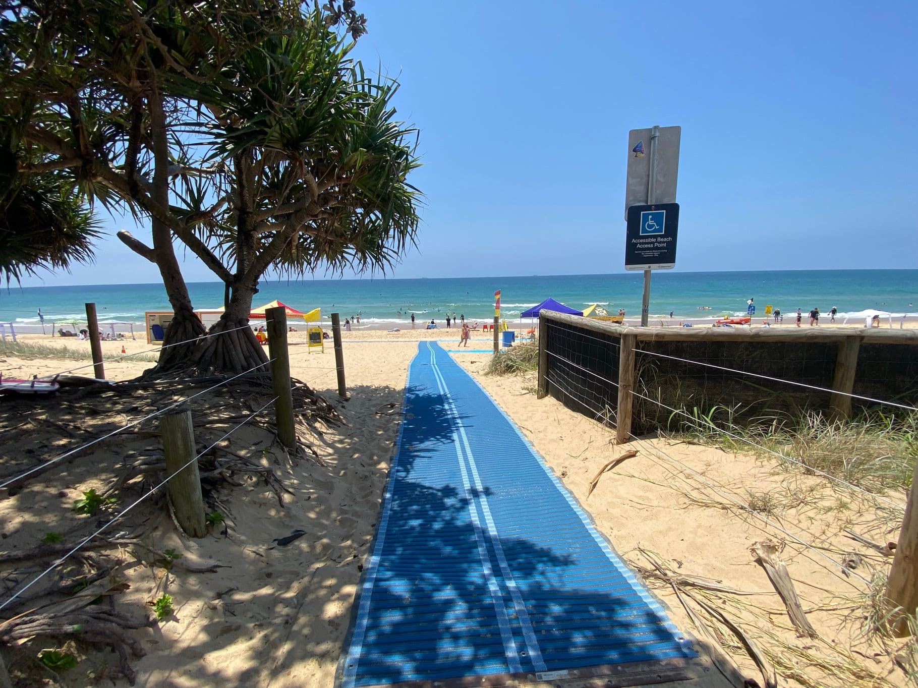 On the left, a large pandanus tree with exposed roots is surrounded by sandy ground and wooden posts connected by wire. On the right, a wooden fence with wire mesh follows the edge of the sandy pathway, with a sign for accessible beach access attached to a post. In the middle, a blue mobility mat stretches across the sand, leading towards the shoreline. The floor consists of sand with patches of vegetation near the edges of the path.
