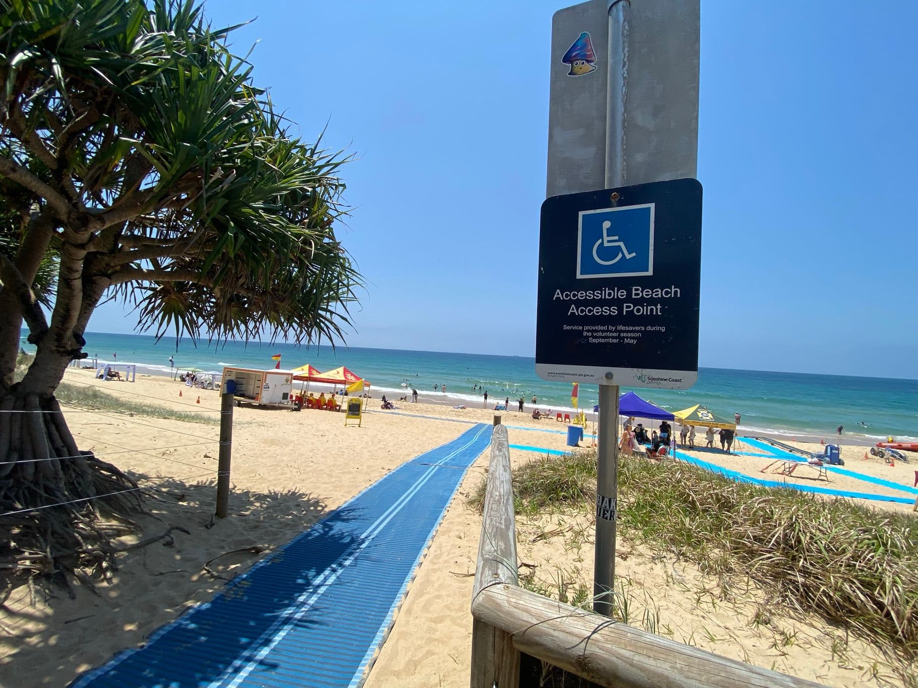 On the left, a large pandanus tree with thick foliage rests on sandy ground near a wooden post with wire fencing. On the right, a blue sign on a metal post reads "Accessible Beach Access Point" above sand and grass. In the middle, a blue mobility mat stretches over the sand towards the beach, with tents and a lifeguard hut visible in the background. The floor is sandy with some grassy patches near the edges of the mat.