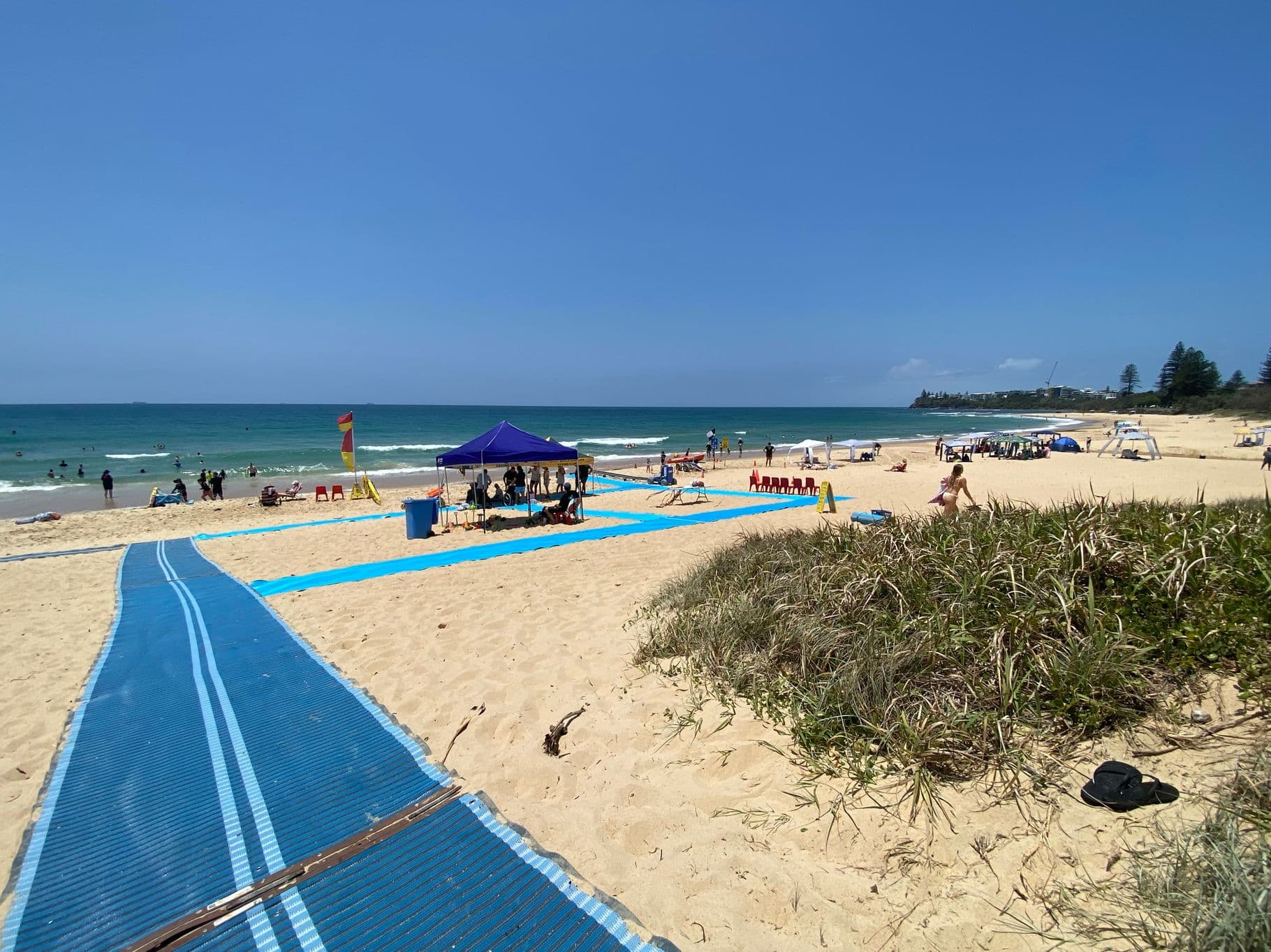 On the left, a blue mobility mat starts near the edge of the image, leading towards the centre across sandy ground. On the right, a patch of grass and low shrubs borders the sand. In the middle, a larger section of the mat runs through the sand towards a purple canopy shelter with people seated underneath. The background includes the ocean, swimmers, a surf lifesaving flag, scattered beach chairs, and other shelters. The floor is sandy with areas of vegetation.