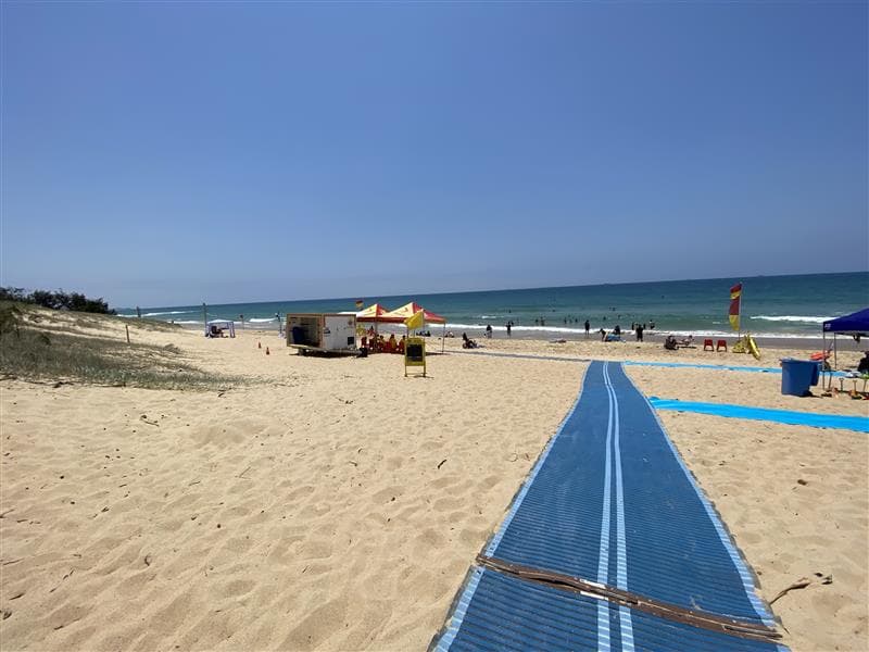 On the left, a grassy dune runs parallel to the sandy beach, with scattered vegetation at its base. On the right, part of a blue mobility mat leads to the beach, next to a blue bin and a smaller mat. In the middle, the main blue mat stretches across the sand towards a yellow and red tent near a trailer. The background includes people swimming in the ocean, a surf lifesaving flag on the right, and additional tents and chairs near the shoreline. The floor is dry sand with footprints.