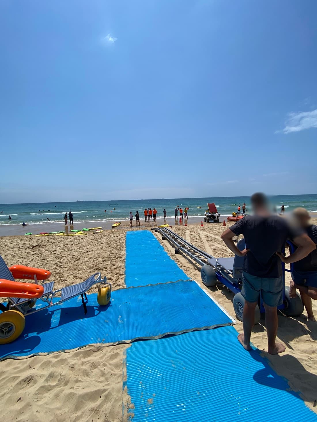 On the left, two beach wheelchairs with orange floats and large yellow wheels rest on a blue mobility mat that stretches across the sandy beach. On the right, a group of people stand near additional equipment, including another beach wheelchair with grey wheels. In the middle, the mat leads down to the shoreline where lifeguards in orange tops stand near the water. The background consists of the ocean with swimmers. The floor is sandy with footprints, and the mat has visible wear.
