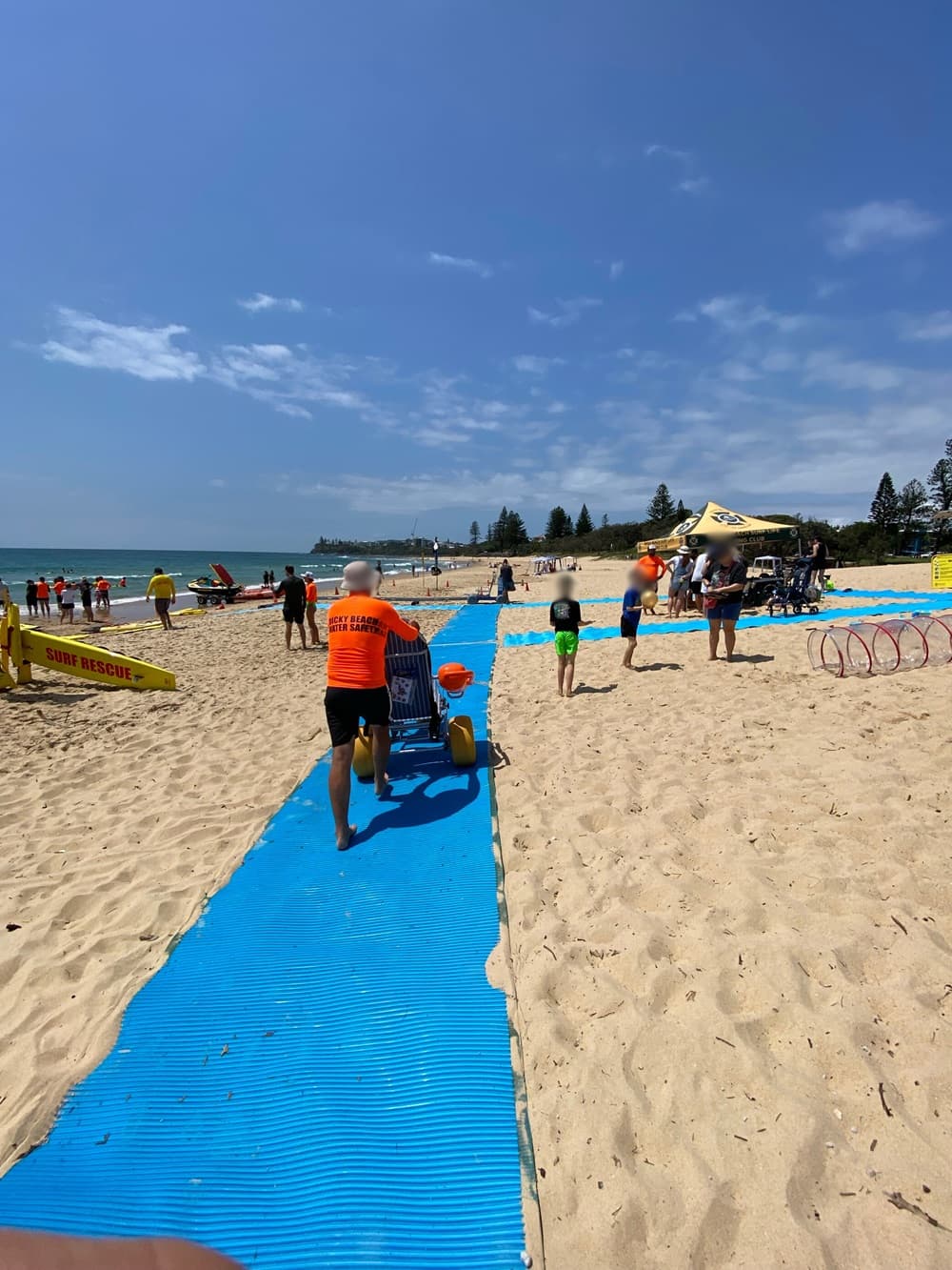 On the left, a lifeguard in an orange shirt pushes a yellow beach wheelchair along a blue mobility mat surrounded by sandy ground. On the right, a group of people gather near a yellow canopy tent featuring surf rescue equipment and hula hoops. In the middle, the mat extends straight through the sand towards the shoreline in the background, where more people stand near the water and lifeguard boats. The floor is sandy with scattered footprints, and the mat forms visible pathways.