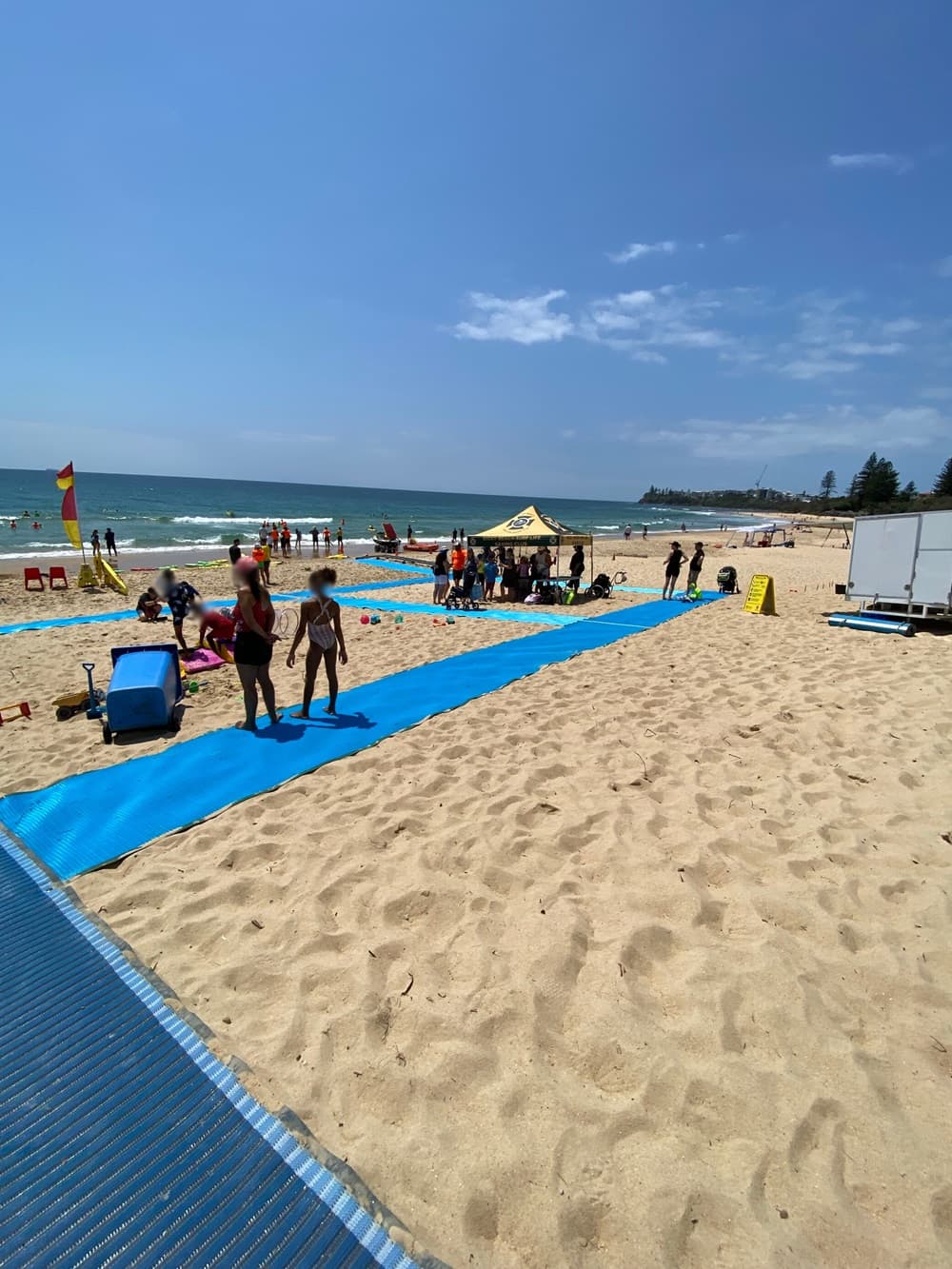 On the left, a blue mobility mat stretches over sandy ground, bordered by a lifeguard flag and beach chairs near the shoreline. On the right, a yellow canopy tent shelters people, with a white trailer and a warning sign further back on the sand. In the middle, another section of the mat extends towards the foreground, bordered by people walking and sitting on the sand. The background includes the ocean edge and distant trees. The floor is sandy, with footprints scattered across the area.