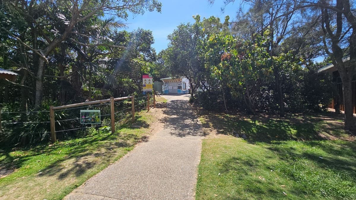 A path runs centre, flanked by grass and dense bushes. To the left, a wooden fence with wire fencing sits along the edge of a trimmed lawn; a small signboard is mounted on the fence. On the right, thick shrubbery and trees create a shaded verge. In the distance, a small building or booth is visible beyond the path, with more greenery framing the scene. The ground is a light-paved footpath; grass borders both sides.