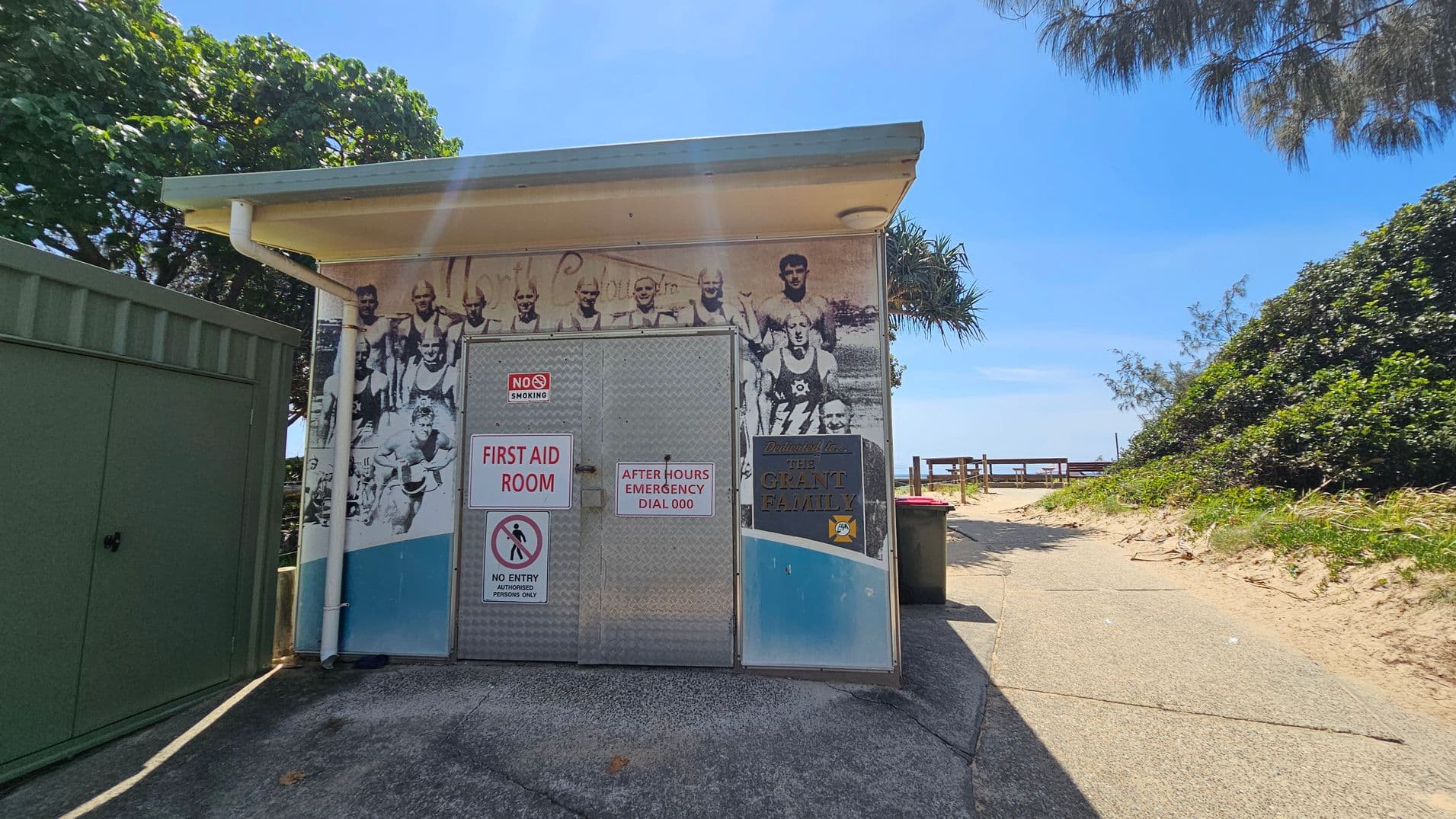 A small structure with a bold metal door in the centre. Left side shows a green utility cabinet and a white pipe running down the wall; right side has a brown rubbish bin. Background features a wooden railing path leading to a sandy slope with bushes on either side. The floor is a grey concrete surface, slightly worn, extending from the front of the door to the path on the right. Ceiling is a flat, light-coloured canopy.