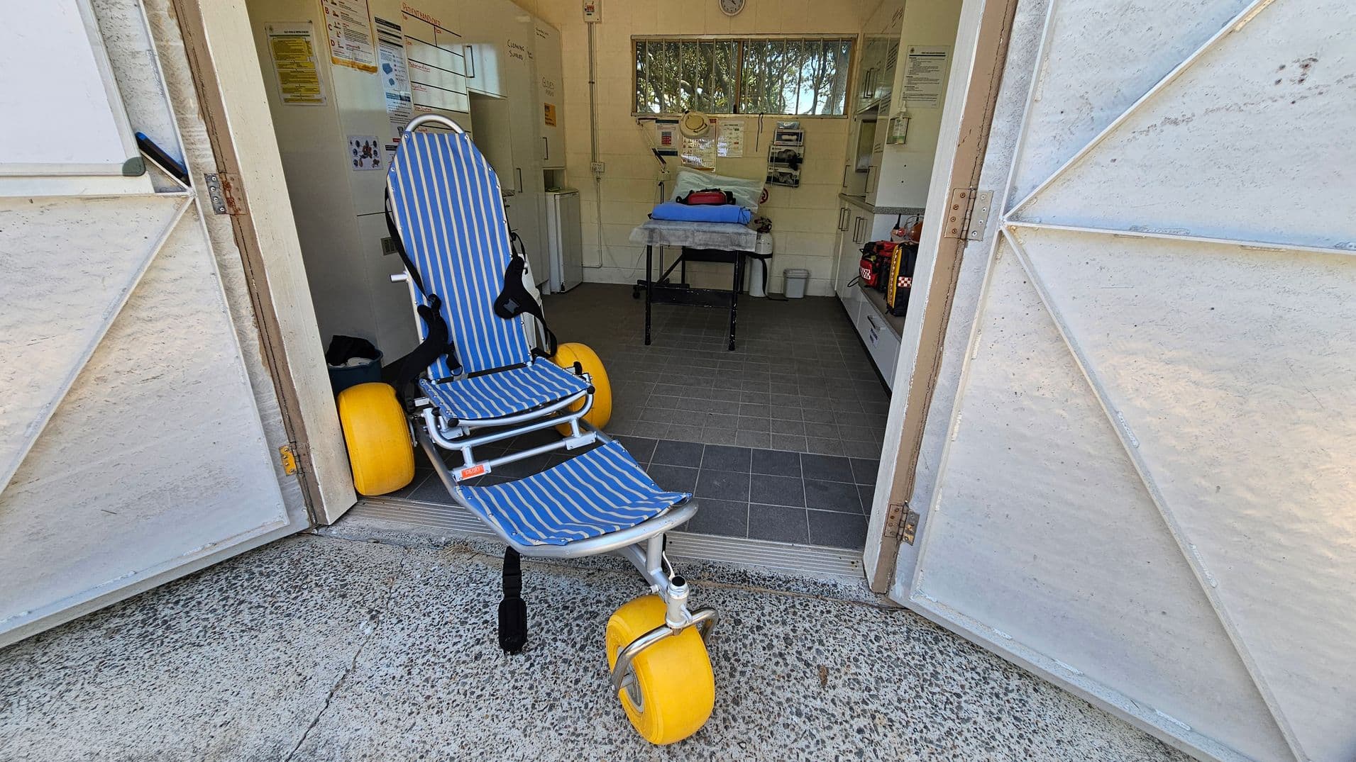 A doorway opens into a small clinic-like room. On the left a striped blue and white reclining chair with large yellow wheels sits just inside the entrance. In the middle inside, a hospital trolley with a blue sheet is visible, along with white cabinets and medical equipment. The floor inside is dark grey tiles; the threshold outside is concrete. Doors are white and open, framing a path that leads to a bright interior.