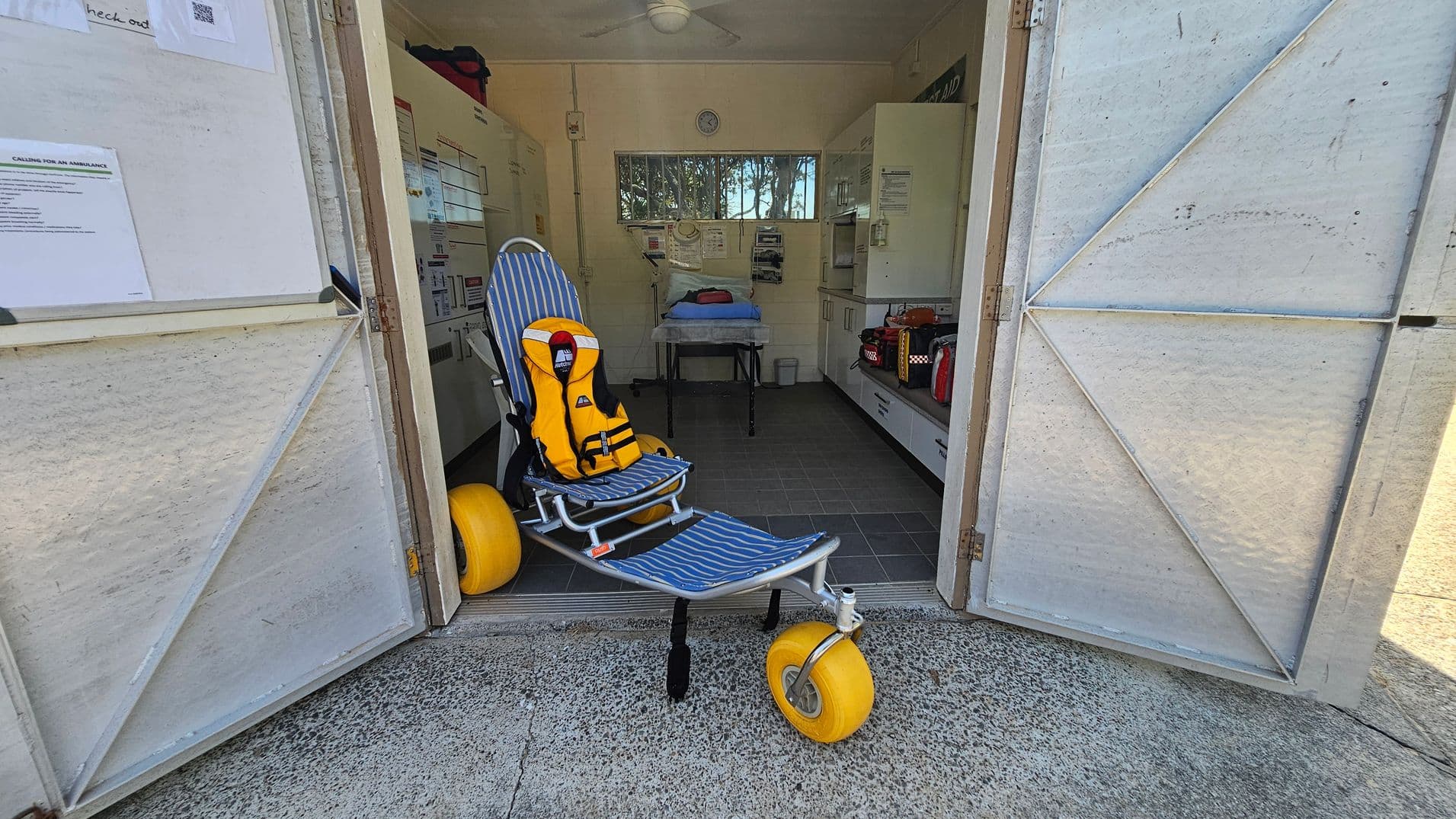 A doorway opens into a small medical room. On the left inside, a tall metal cabinet lines the wall. Center is a blue-and-white striped reclining chair with a bright yellow life jacket secured on it. A single large yellow wheel sits at the chair’s rear. In the middle/background, a hospital trolley with a blue sheet stands near a workbench and white cupboards. The floor is dark grey tiles inside; the outer threshold is rough concrete.