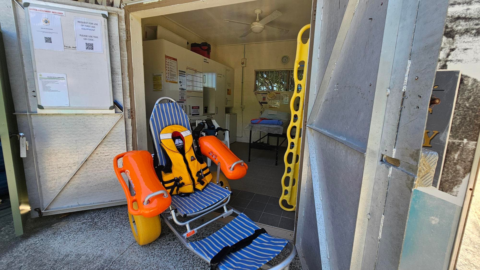 A doorway opens into a small medical room. On the left outside, a white utility box is mounted on a light wall. Inside near the threshold, a blue-and-white striped reclining chair with orange forearm supports sits on the concrete floor, beside a yellow life-raft seat. A second orange seat is attached to the chair. Inside, a grey tiled floor leads to white cupboards and a hospital bed in the background.
