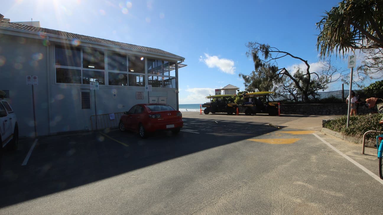 A paved area with parking lines is in the foreground. On the left, a grey building with large windows and a tiled roof stands beside a red car and part of a white vehicle. On the right, trees and bushes border the space, with a bicycle rack and a sign for parking visible near the edge. In the middle, two yellow utility vehicles are parked, and in the background, a small gazebo-like structure overlooks the sea surrounded by sandy ground and sparse trees.