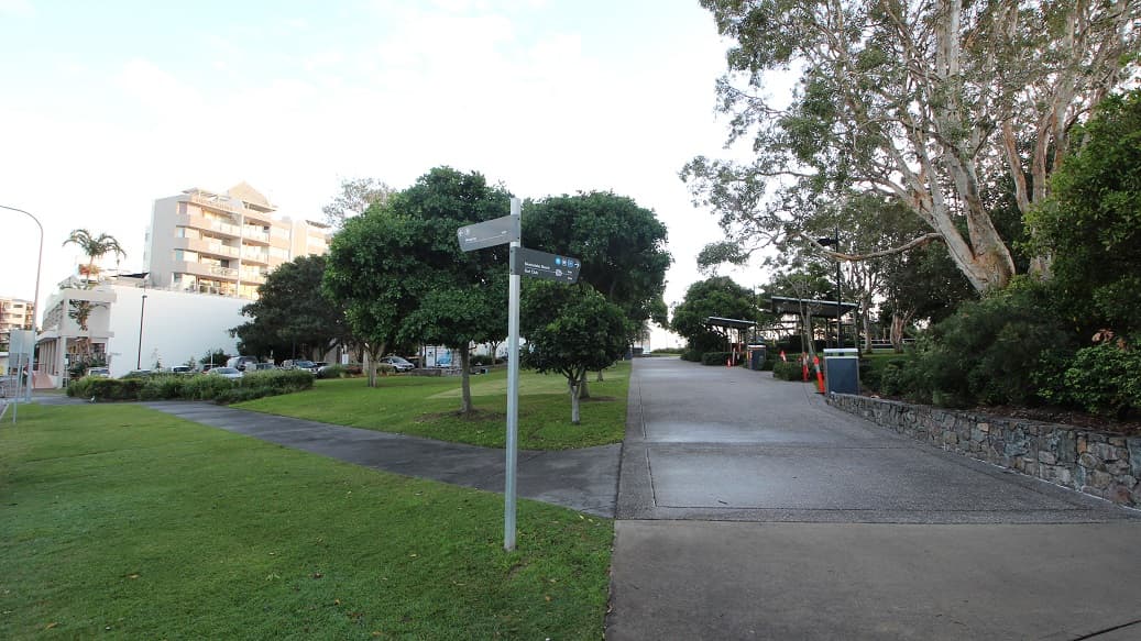 A paved path runs from the foreground into the middle, bordered by grass on the left and a low stone wall with trees on the right. On the left, a modern apartment building and parked cars are partly visible near clusters of trees. On the right, large trees with overhanging branches and shaded seating areas with structures appear alongside a bin. In the background, the path continues, flanked by trees and landscaping. The floor is a mix of grass, pavement, and concrete.