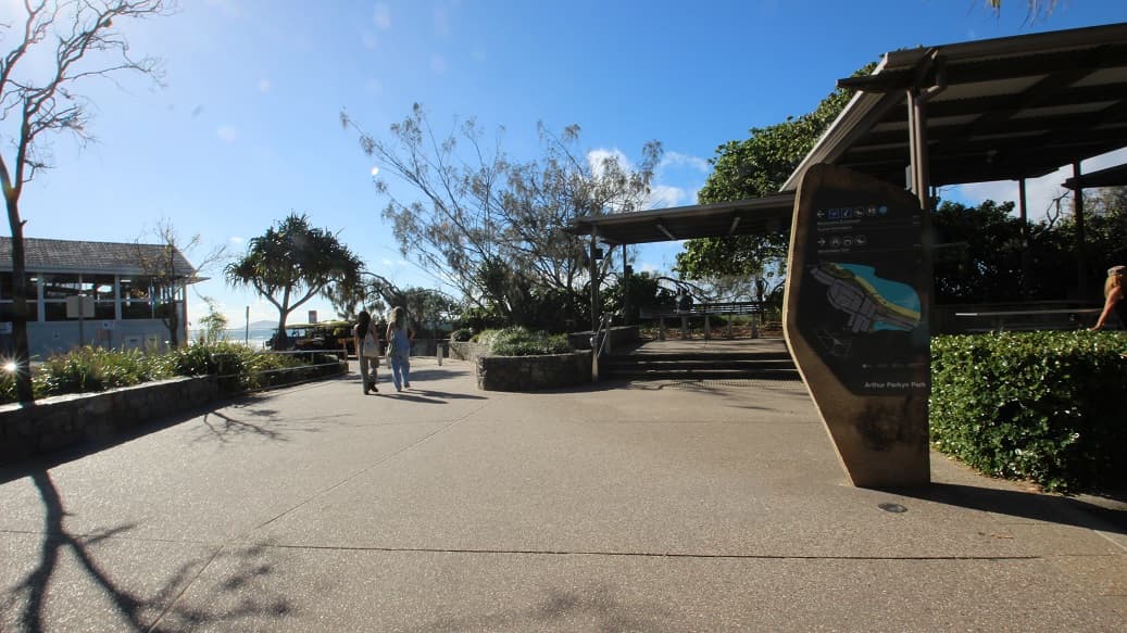 A paved walkway stretches across the foreground and middle, with a large stone map display on the right in front of green hedges. On the left, there’s a low stone wall bordering plants, and a building with a tiled roof sits further back. In the middle, two people walk near sparsely leafed trees, with benches and planters on either side. To the right, a covered seating area is elevated by steps, partially shaded by foliage. The floor is textured concrete, and canopy structures form the ceiling.