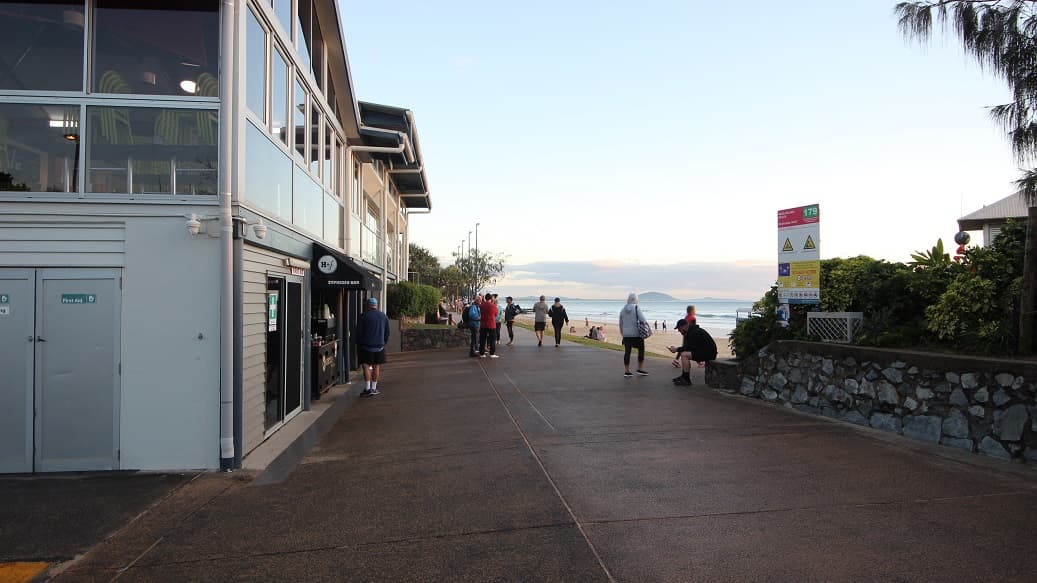 On the left, a two-storey modern building with glass windows, a black awning reading "Espresso Bar," and grey siding. Below are doors and a service counter. On the right, a stone wall with plants and safety signs. In the middle, a paved walkway leads towards the background, where people are walking or sitting near the beach. The floor is a brown-toned textured pavement.