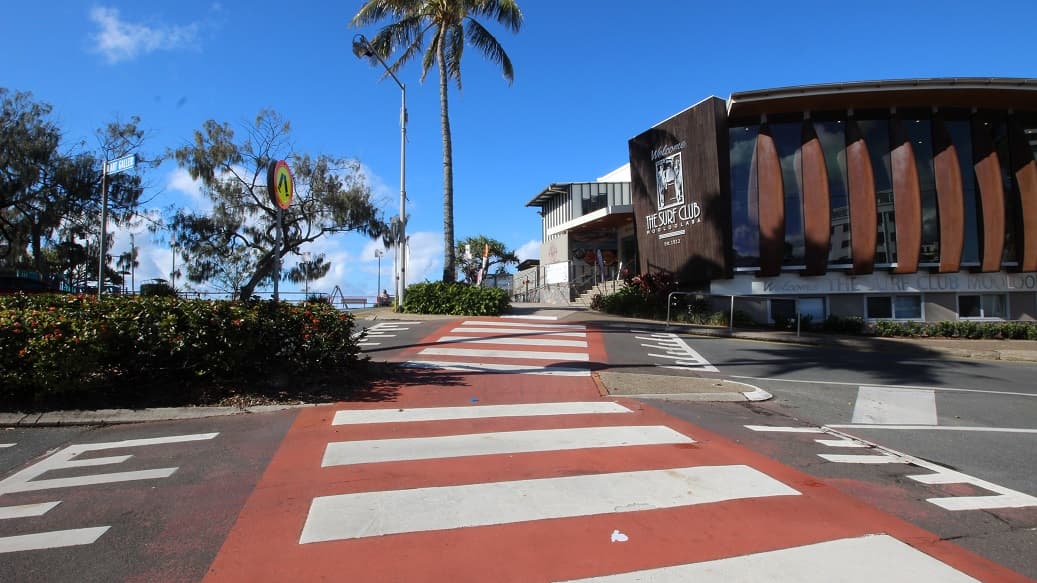 A zebra crossing with alternating red and white painted sections spans the foreground over a paved road. On the left, a hedge with flowering plants lines the side near a circular pedestrian crossing sign and tall trees. On the right, a modern surf club building with wooden panel accents and large windows rises slightly above street level, bordered by steps and greenery. In the middle, a palm tree stands next to a pathway leading towards distant railings. The floor is painted and paved.