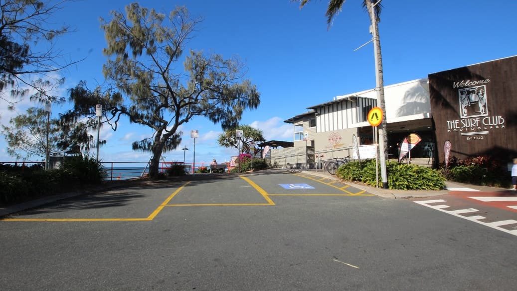 A paved road with yellow markings slopes upward into the background, flanked by greenery. On the left, tall trees with sparse foliage stand near a metal railing that borders the edge of a pedestrian walkway. On the right, a building with a wooden front reading “The Surf Club” is surrounded by plants, bicycles, and signage. In the middle, the road leads to a pathway lined with a tree, pink flowers, and benches overlooking the water. The floor is paved, and the ceiling is open-air.