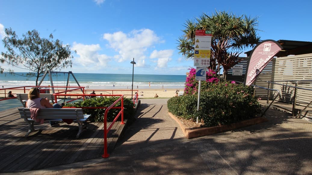 On the left, a wooden seating area with benches overlooks a sandy beach, bordered by a red metal barrier and greenery. On the right, a garden bed with pink flowers and a tall pandanus tree sits beside a wooden building with horizontal slats and a banner that reads “Welcome.” In the middle, a concrete pathway leads down to the beach, with safety signs and a lamp post near the steps. The floor is a mix of wooden decking, concrete, and sand.