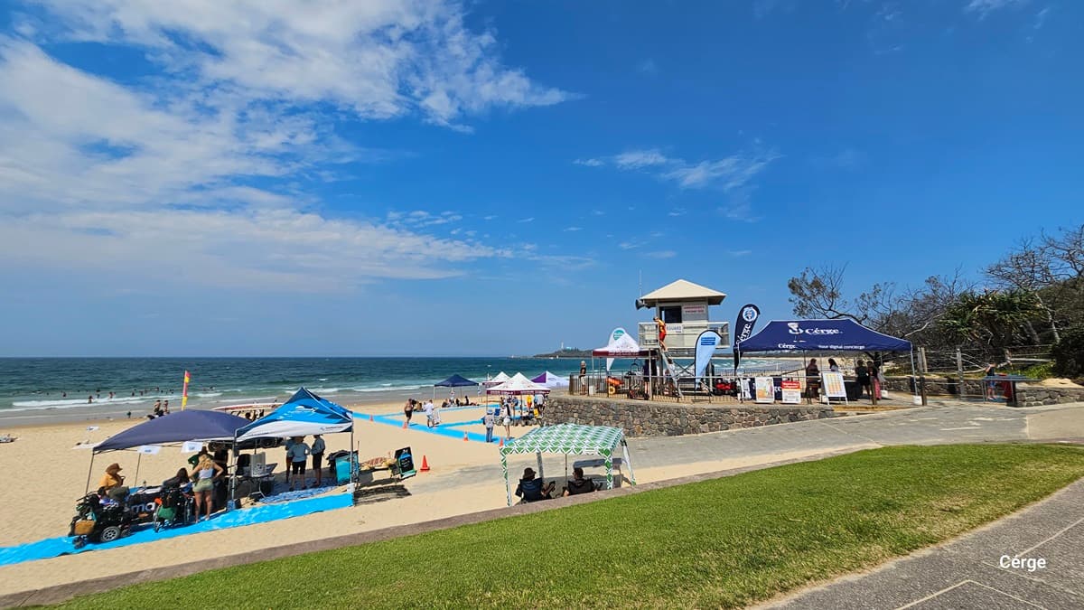 On the left, a sandy beach with swimmers and a flagged surf zone is bordered by small blue tents and event stalls on a bright blue path. In the middle, a stone retaining wall supports a lifeguard tower surrounded by flags, banners, and more tents. On the right, a paved footpath runs along grass and trees, with additional stalls and railings near the edge. The floor is sand, pavement, and grass. There is no visible ceiling.