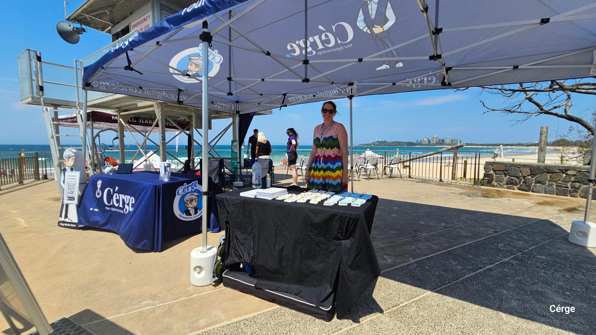 A large canopy is set up in the foreground, covering a table on the right with a black tablecloth displaying promotional items. A person stands behind it. On the left, another table covered with a blue cloth displaying “Cérge” branding sits near a cardboard cutout and a lifeguard tower on a sandy surface. In the background, a low metal railing overlooks the beach and ocean, with a stone wall on the far right. The floor is a mix of concrete and sand. The ceiling is the underside of the canopy.