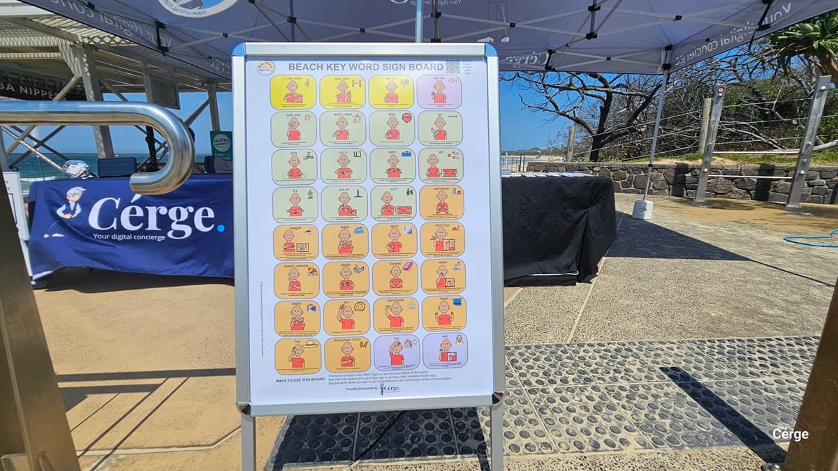 On the left, a blue tablecloth with the “Cérge” logo covers a table under a canopy structure with visible metal beams. On the right, a black tablecloth covers another table, set behind a roped railing near a stone wall and greenery. In the middle, a freestanding signboard displays illustrated “Beach Key Word” symbols. The floor consists of textured concrete and rubber mats. The ceiling is formed by angled canopy panels.