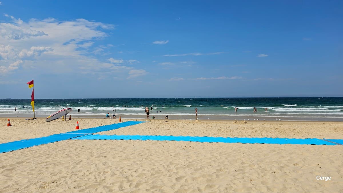 On the left, a red and yellow flag stands near a tilted surfboard resting upright on the sand with a few orange traffic cones nearby. On the right, small groups of people are scattered along the shoreline and in the water. In the middle, a wide blue mat creates a pathway across the sandy beach, leading towards the waves. The foreground is covered in golden sand, and the background features the ocean with small waves breaking near swimmers.