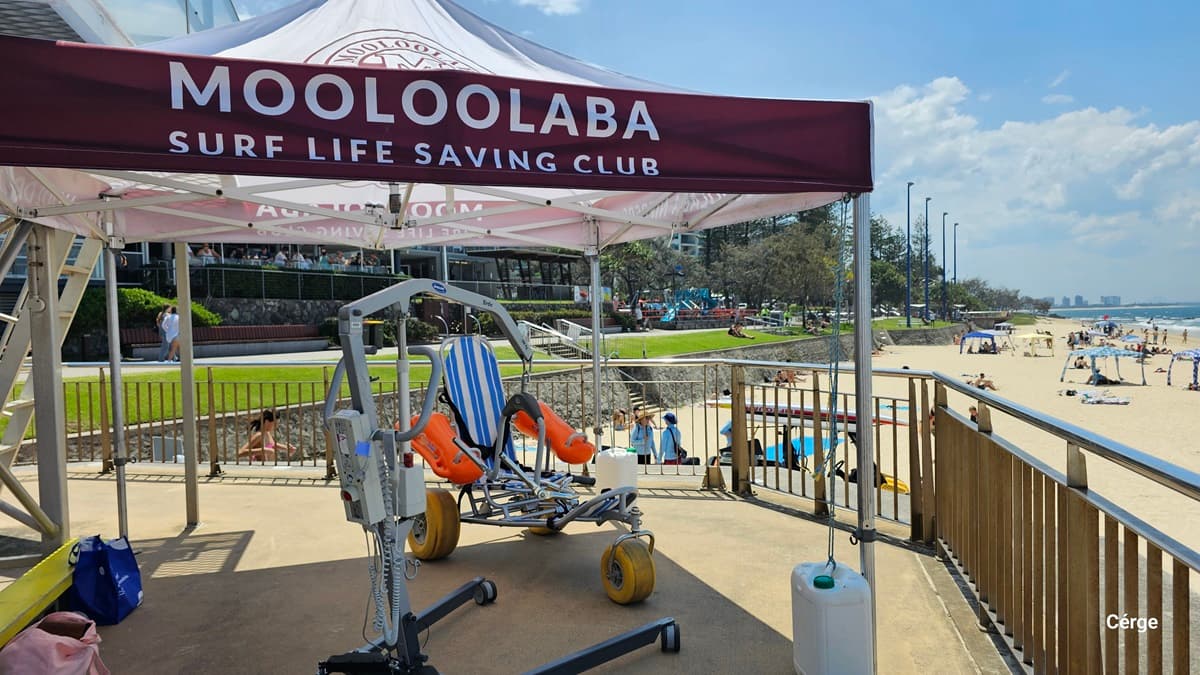 Under a maroon and white canopy labelled “Mooloolaba Surf Life Saving Club,” a mobility beach chair with orange floatation devices rests on a smooth concrete platform. On the left, a blue bag sits near the canopy’s pole. On the right, a wooden railing overlooks a sandy beach with scattered umbrellas and people. In the background, a grassy park area with pathways, benches, and trees is visible above a stone retaining wall. The floor is concrete, and the ceiling is the canopy.