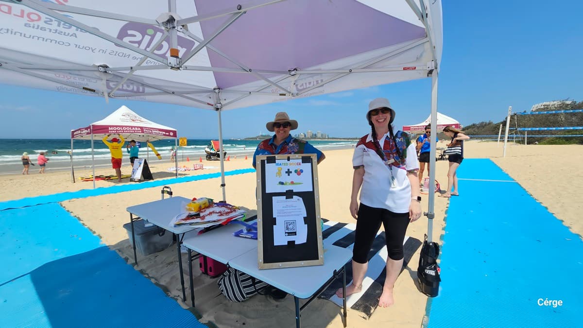 Under a white canopy, two women wearing hats stand on a striped mat behind a table. On the left, a blackboard easel with event information is positioned near colourful items spread across the table. In the background, another canopy with “Mooloolaba Surf Life Saving Club” branding is near a lifeguard in yellow and red. On the right, a bright blue mat stretches across the sandy beach toward volleyball nets. The floor is sand, and the ceiling features the canopy's framework.