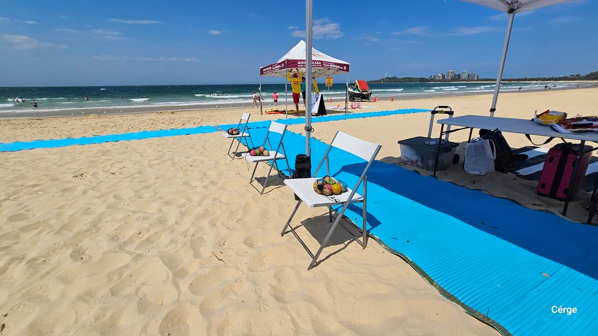 On the left, a long blue mat stretches across the sandy beach, leading toward the water where people are swimming. In the middle, three white folding chairs with baskets of fruit sit on the mat, partially shaded by a white canopy. On the right, a table with containers, bags, and a pink suitcase is positioned near the mat. In the background, a maroon and white tent with lifeguards and a beach mobility chair is visible. The floor is sand, and the canopy overhead forms the ceiling.