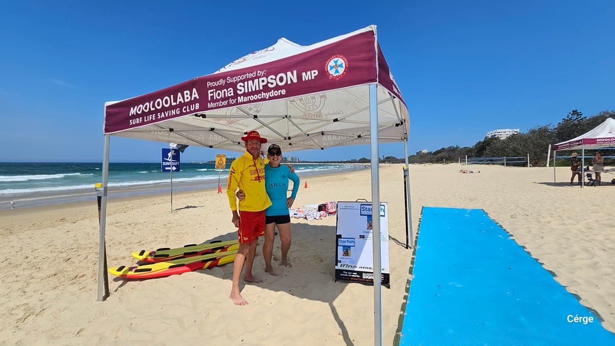 On the left, yellow and red rescue boards lie on the sand near a pole supporting a maroon canopy that reads “Mooloolaba Surf Life Saving Club.” In the middle, two people stand under the canopy beside an easel with information boards. On the right, a bright blue mat stretches across the sand, leading toward two additional canopies in the background. The floor is sand, and the ceiling is the underside of the canopy with visible support beams.