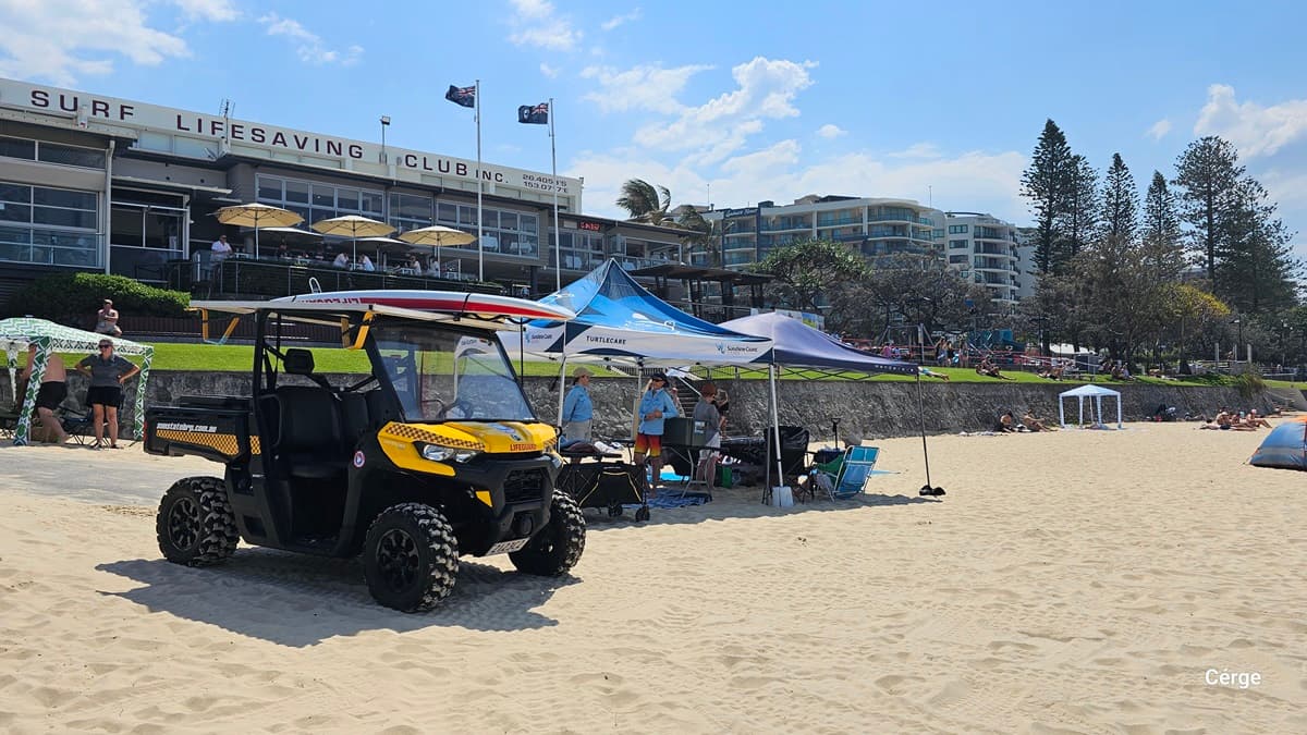 On the left, a black and yellow lifeguard buggy with thick tyres is parked on the sand. In the middle, two canopies, one blue and one white, provide shade for several people standing and sitting on beach chairs beside tables. On the right, beachgoers sit under smaller structures or relax on the sand. In the background, a surf lifesaving club building with umbrellas and flags overlooks the area, with apartment buildings and tall trees visible further to the right. The floor is sand.