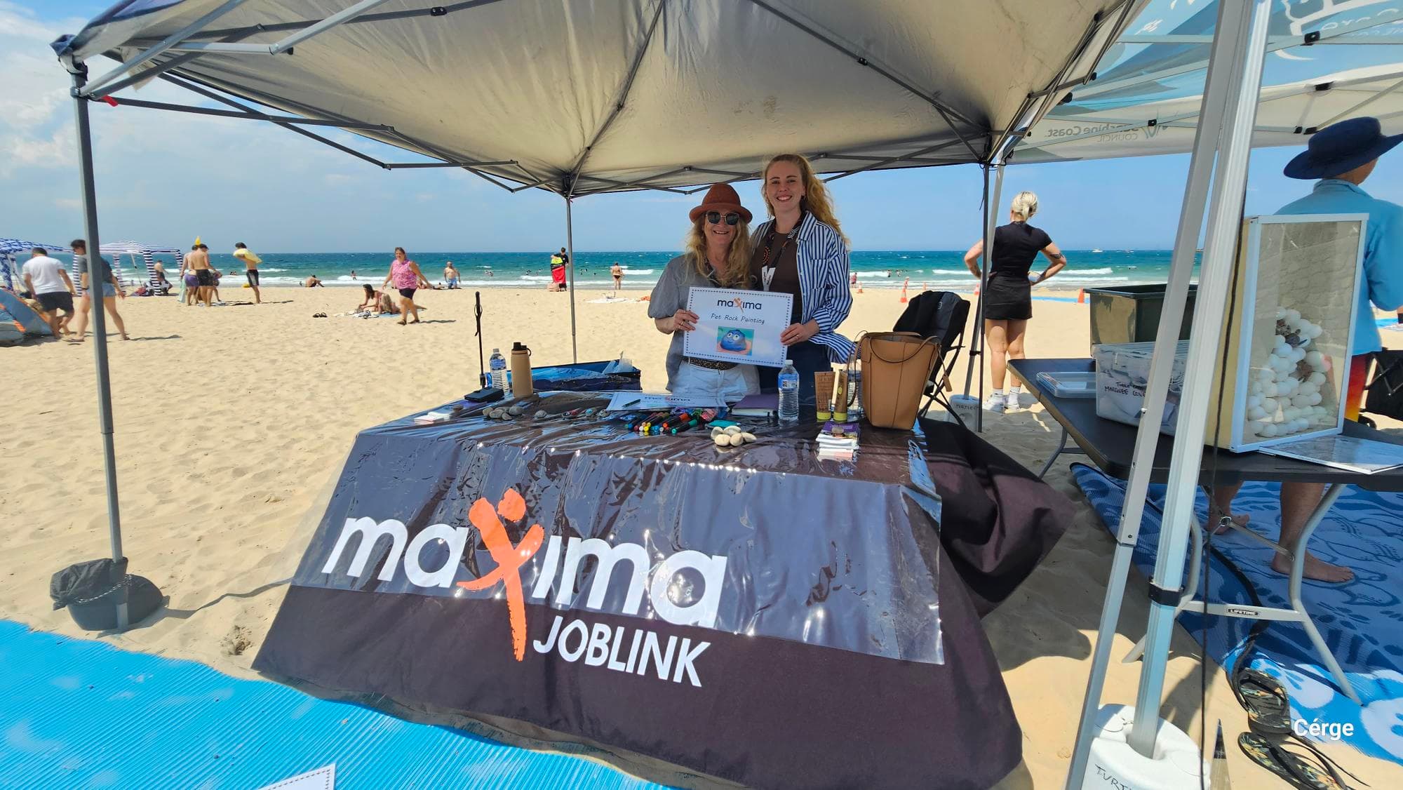 Under a grey canopy, two women stand behind a table covered with a black cloth displaying the "Maxima Joblink" logo. On the left, the sandy beach extends, with scattered people walking and sitting near the water. On the right, a table holds a clear display box with white balls, and a woman in a blue hat stands nearby. In the foreground, a textured blue mat lies on the sand. The ceiling is the underside of the canopy with visible metal supports.