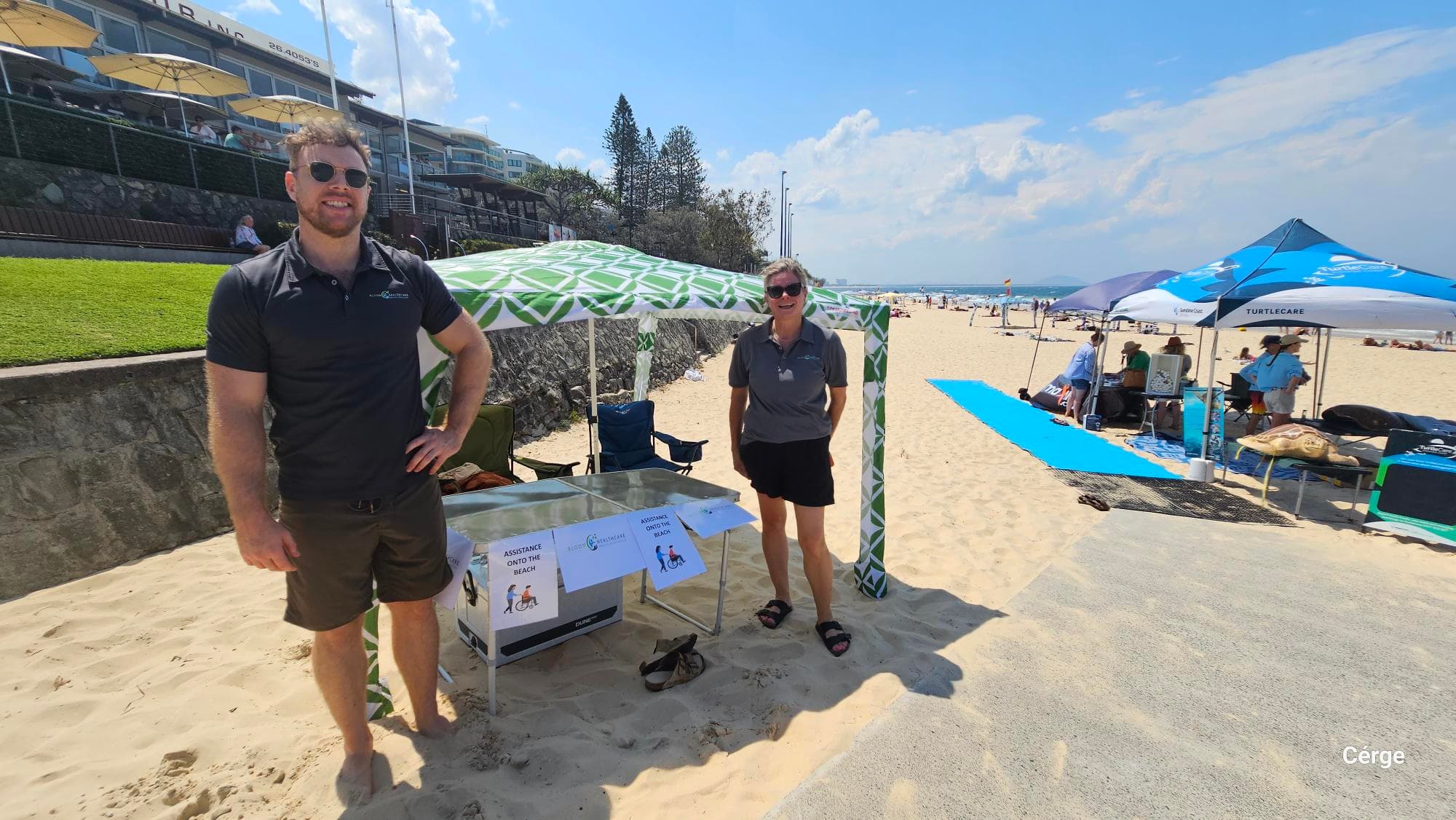 On the left, a man stands on the sand next to a short stone retaining wall with grass above it, leading to a building with umbrellas. In the middle, a green and white patterned canopy shades a table holding brochures and signs reading “Assistance on the Beach.” On the right, a woman stands beside the canopy, and further right, a blue beach mat leads to additional canopies and chairs. The floor is sandy, and the ceiling is formed by the canopy overhead.