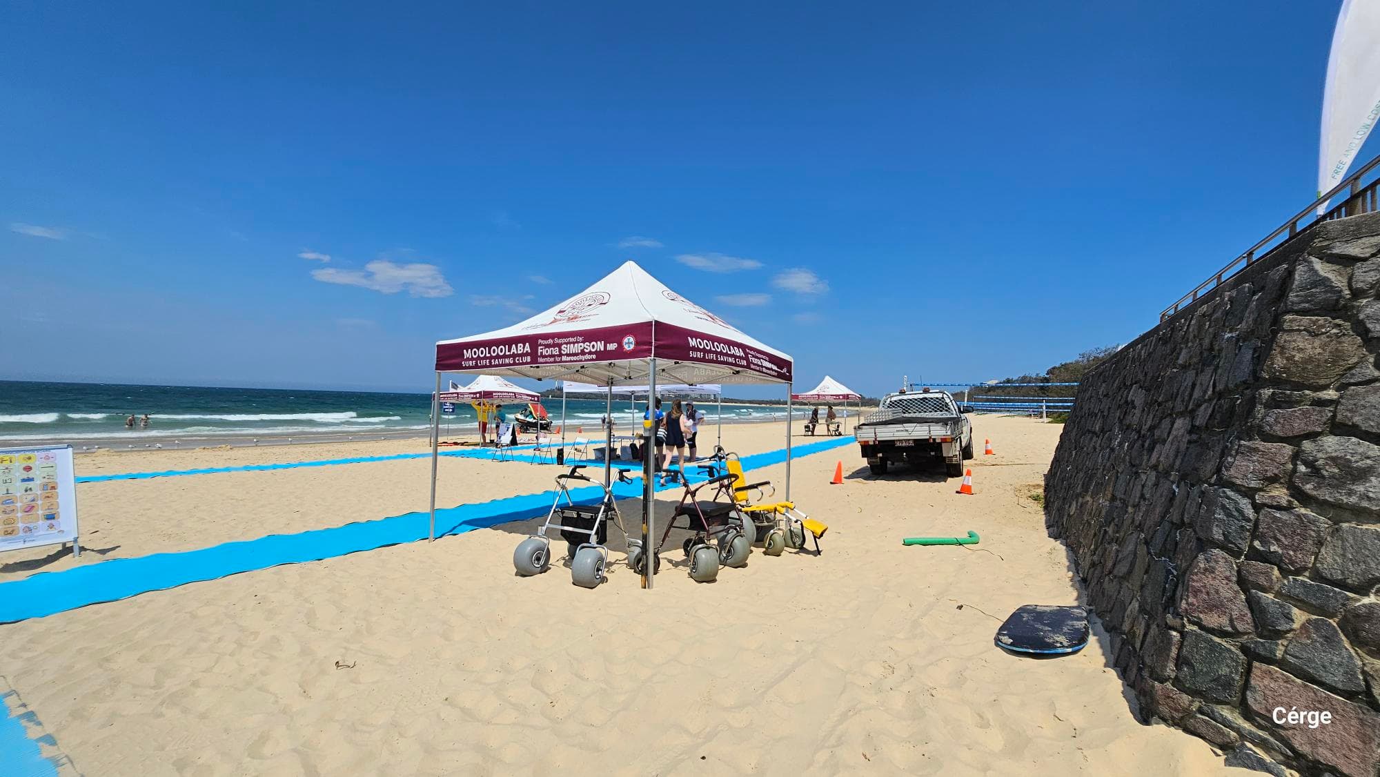 On the left, two blue mats stretch across the sandy beach towards the water, with a bright information board nearby. In the middle, a white canopy marked “Mooloolaba Surf Life Saving Club” shades beach-access wheelchairs, chairs, and several people walking. On the right, a parked utility vehicle is near traffic cones, while a stone retaining wall runs along the edge of the sand. The floor is sandy, and the canopy overhead forms the ceiling in the middle section.