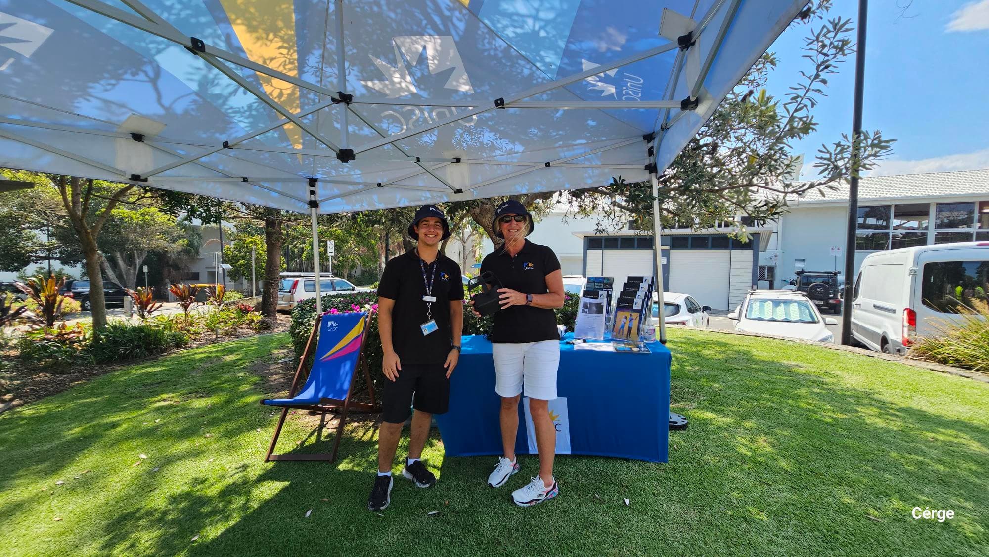 On the left, trees and landscaped bushes border a footpath near parked cars. In the middle, two people in black shirts and hats stand under a white canopy beside a table draped in a blue cloth displaying promotional materials and brochures. On the right, more trees, a white van, and a building with large windows are visible. The floor is neatly trimmed grass, and the ceiling is the inner frame of the white canopy.